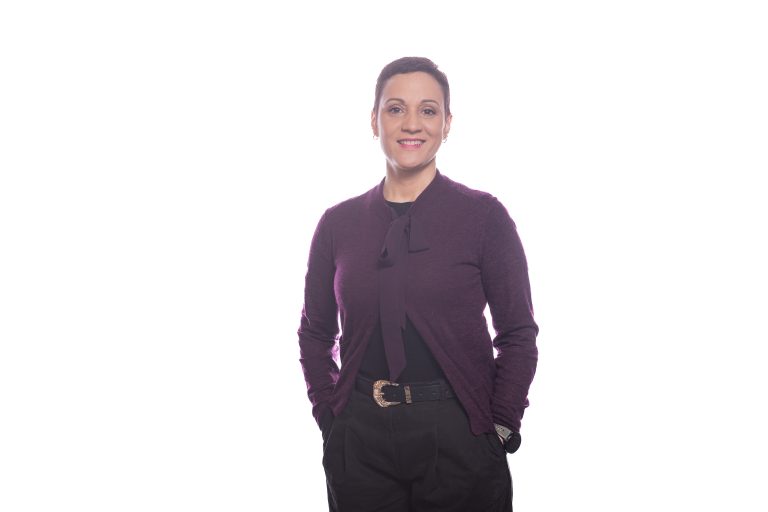 Retrato plano americano de Remy de pie, de frente, con un ligero escorzo y las manos en los bolsillos. Lleva una chaqueta lila/morada y pantalones negros, con fondo blanco.